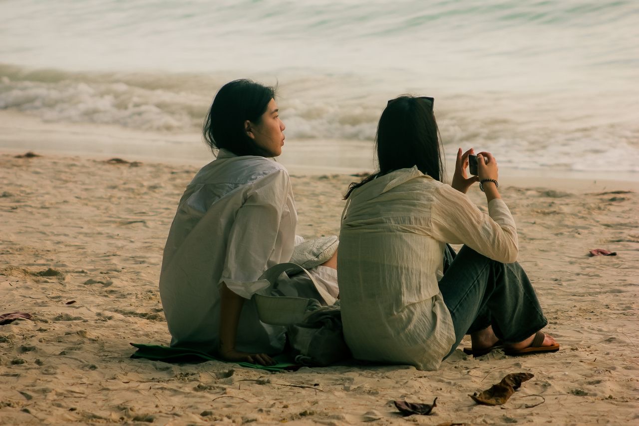 Two friends sitting together on the sand, looking out at the sea